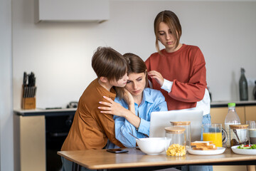Obraz na płótnie Canvas Mom having problems at work working online. Teen children supported her in stressful moment. Boy and girl hug mother working on laptop sitting at table on kitchen at home. Combining family and career.