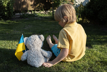 boy and his imaginary friend, a toy bear with a Ukrainian flag, are sitting on the grass with their...