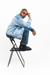 young and handsome Afro-American man in cool denim clothes sitting on the back of the chair and looking serious, studio shot vertical. High quality photo