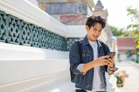 Portrait of Asian male traveler using a mobile phone on sidewalk of buddhist temple on street in Bangkok, Thailand, Southeast Asia - smart phone and internet for traveling concept - Powered by Adobe