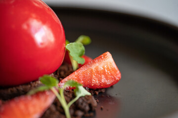 Strawberry dessert detail. Sphere shaped curd cake with strawberries and brownie crumbs. Red dessert on the black plate. Selective focus.