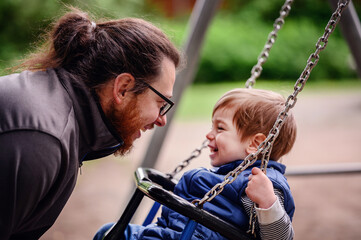 Obraz premium Little toddler boy happily rides on swings and playing with tall long haired man in glasses