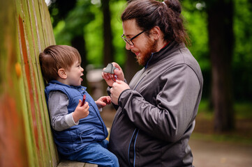 Tall long dark haired man with ponytail and glasses stands near wooden fence with little toddler boy