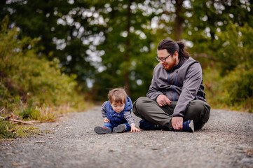 Fototapeta premium Young long hair bearded man in glasses sitting with little toddler boy on road playing with stones
