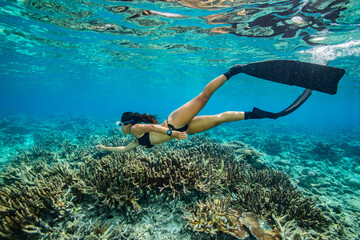 A young woman in bikini free dives above a coral reef in clear tropical water	