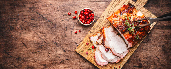 Baked pork loin on rustic wooden cutting board with spices, herbs and cranberries. Wood kitchen table background, top view banner with copy space - obrazy, fototapety, plakaty