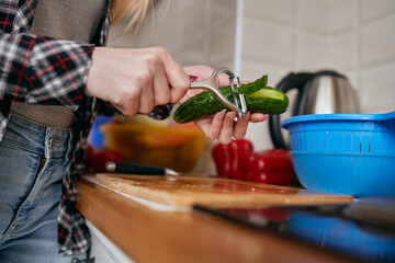 Female person peeling off a cucumber skin with a peeler tool. Housewife preparing a fresh vegetarian salad with low carb vegetables in a home kitchen