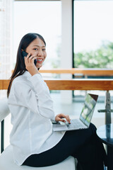 Obraz premium A young Asian self employed woman showing a smiling face and happiness while using a computer and using smartphone mobile talking with business at a cafe shop. self-employed, freelance, relaxation.