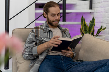 Man reading book while sitting on cough at home, enjoying interesting novel.