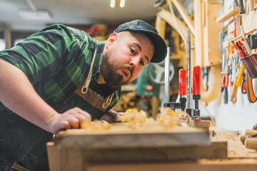Focused male carpenter blowing wood shavings off the wooden plank in a workshop. High quality photo