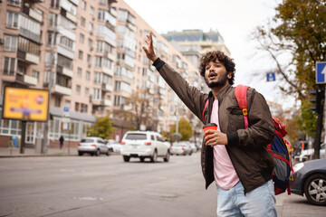 Indian backpacker with coffee to go catching taxi near road on city street