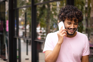 Smiling young indian man in pink t-shirt talking on smartphone while walking near building on city street 