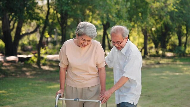 Asian senior husband take care to wife having knee pain while walking with walker stick in the park, Physical therapy in outdoor concept