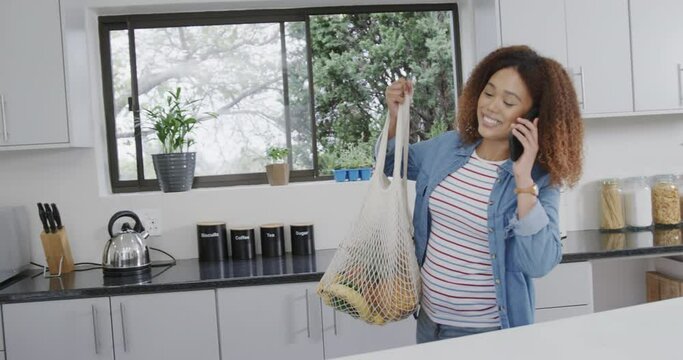 Happy biracial woman talking on phone putting down bags of grocery shopping in kitchen, slow motion