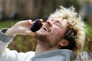 A bearded blond man is having a lot of fun talking on the phone in the park on a bench.