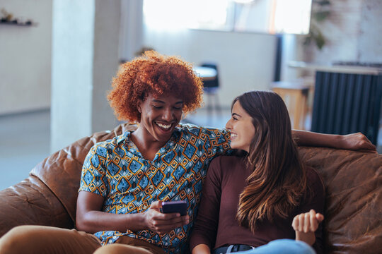 Two young multiracial people sitting on couch in living room and bonding while using phone.
