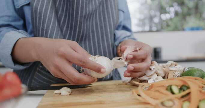Midsection of biracial woman in apron preparing meal, peeling garlic in kitchen, slow motion