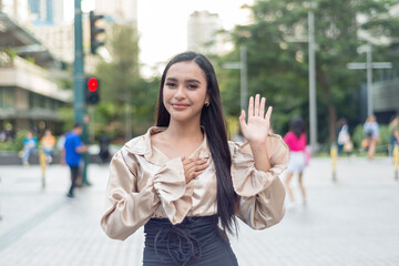 Fototapeta premium A pretty woman standing outside wearing a light tan silk blouse in an oath taking pose while smiling at the camera. Trees, building, people and traffic light are shown in the background.