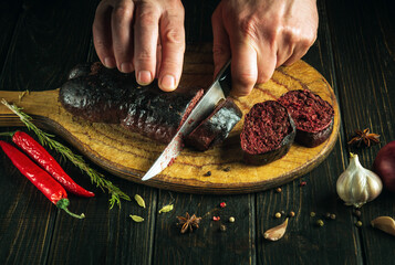 The chef cuts blood sausage on kitchen cutting board with knife in his hand. The concept of cooking...