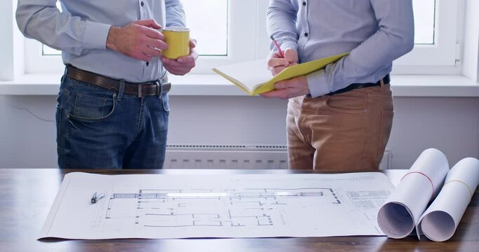 Investor with yellow cup in hands explains to engineer all requests about new building project. Engineer takes notes while stands near table with drawings