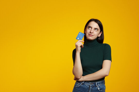 Thoughtful young woman wearing green shirt holding plastic credit card near face and looking at free empty space, isolated over yellow background
