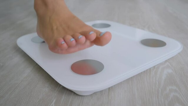 Female bare feet stepping on white digital floor scales - woman weighing herself at home: close up, low angle ground view. Measuring weight, control, wellness and diet concept