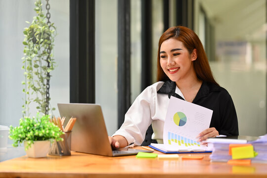 Image of cheerful asian female office worker reading email, using laptop on wooden desk. Business and technology concept