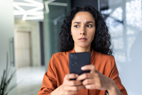 Upset thinking business woman close up inside office, young hispanic woman reading bad news on phone online, business woman at workplace holding smartphone typing text message.