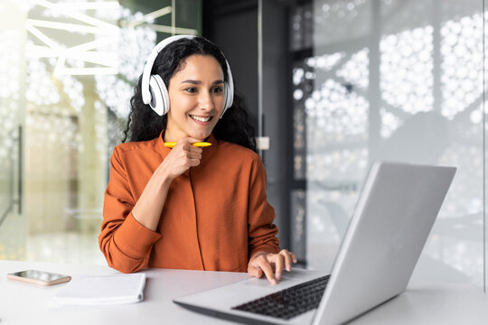 Young curvy businesswoman with headphones listening to online course and audio podcast female worker inside office working with laptop, successful hispanic woman smiling satisfied with work.