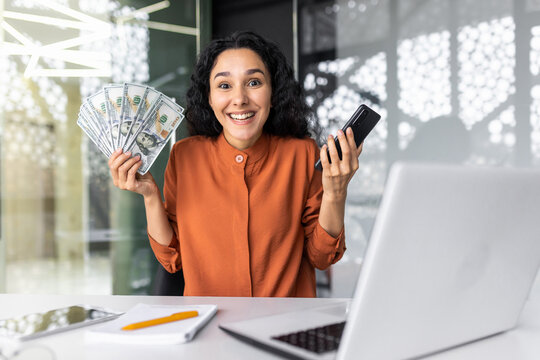 Successful hispanic woman looking at camera and smiling, business woman holding bank credit card and money cash dollars, portrait of successful woman about , office workplace with laptop