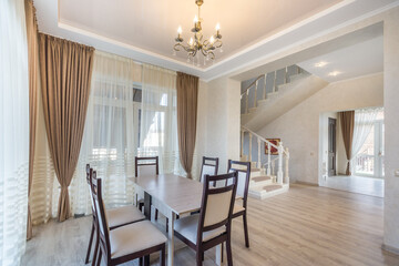 The interior of a country house with a modern design. Elegant chandelier over the dining table, windows with beautiful curtains, stairs to the second floor.