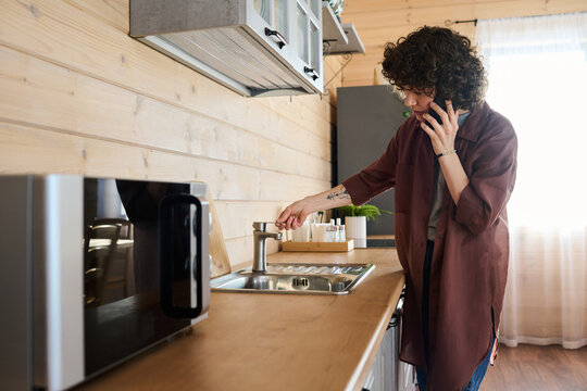 Young housewife with smartphone by ear calling repair service while standing by counter with sink and broken tap in the kitchen