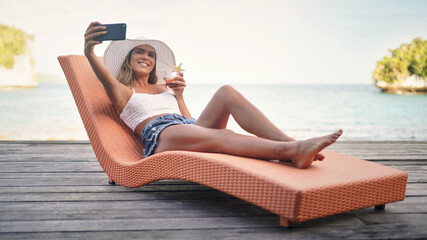 Selfie, travel and woman with water, summer and happiness on a sun lounger, cocktail and tropical island. Female person, girl or happy tourist on a break, social media and drink to relax and cheerful