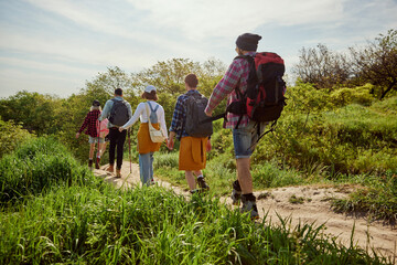 Fototapeta na wymiar Young people, group of friends in comfortable clothes with backpacks walking in forest, going hiking on warm summer day. Concept of active lifestyle, nature, sport and hobby, friendship
