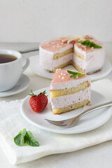 A creamy strawberry slice of cake rests on a white saucer. A cup of tea in the background. Strawberries on the table. Light background