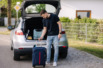Handsome bearded man stand with suitcase in front of open car trunk. Travel road trip. Holiday on summer vacation concept