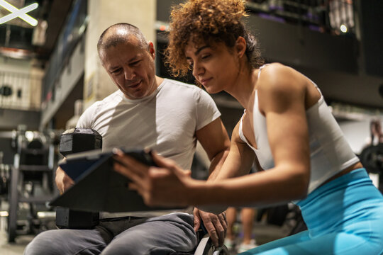 A determined individual in a wheelchair, working out in the gym with the guidance of a dedicated female fitness trainer who using a tablet to show a weekly workout plan.
