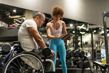 A determined individual in a wheelchair, working out in the gym with the guidance of a dedicated female fitness trainer who using a tablet to show a weekly workout plan.