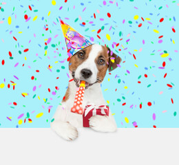 Jack russell terrier puppy wearing a party cap blows into party horn and holds gift box above empty white banner - obrazy, fototapety, plakaty