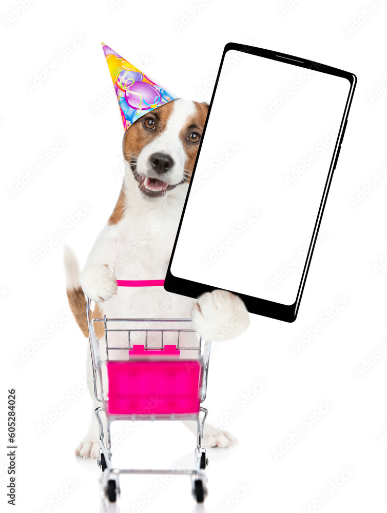 Poster jack russell terrier puppy wearing party cap pushing a shopping cart and holds big smartphone with w