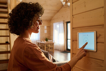 Fototapeta na wymiar Side view of mature brunette woman pointing at screen of alarm system while choosing or adjusting settings on interactive panel