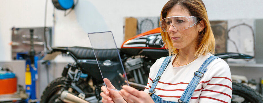 Concentrated female mechanic with security glasses holding transparent tablet in front of motorcycle on garage