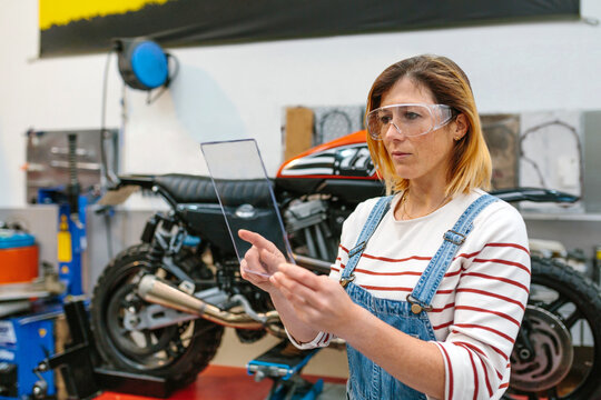 Concentrated female mechanic with security glasses touching transparent tablet in front of motorcycle on garage
