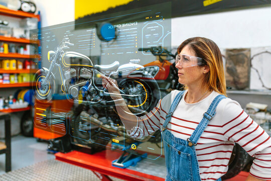 Young female mechanic with glasses touching hud panel screen with augmented reality hologram to review motorcycle on factory. Futuristic user interface concept.