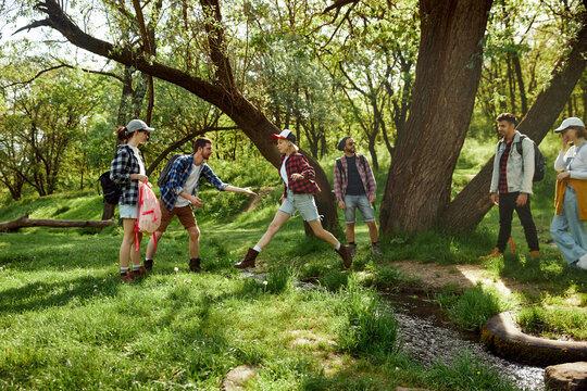 Young people, men and women, friends going hiking, walking in forest, jumping over river on warm day. Concept of active lifestyle, nature, sport and hobby, friendship, leisure time, fun