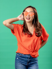 A portrait of an Asian woman wearing an orange shirt and glasses, feeling dizzy and holding her head with one hand, isolated on a green background.