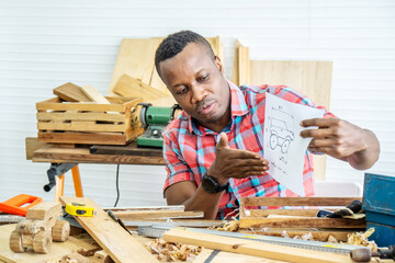 Young african american male carpenter sitting at table showing model car wooden and drawing paper through tablet while video online chatting with customer or teaching online in factory