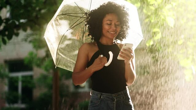 Curly young african woman hold smartphone scrolling social media texting browsing online under umbrella while rain drops falling at sunset park Pretty student relax enjoying great rainy day outdoors