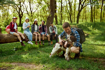 Young man playing with dog. Group of young people, friends resting in forest, going hiking on warm...