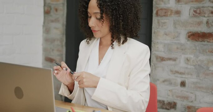 Thoughtful biracial businesswoman sitting at desk holding pen and using laptop, in slow motion